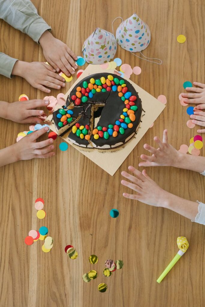 Top view of children's hands reaching for a chocolate birthday cake with colorful candies.